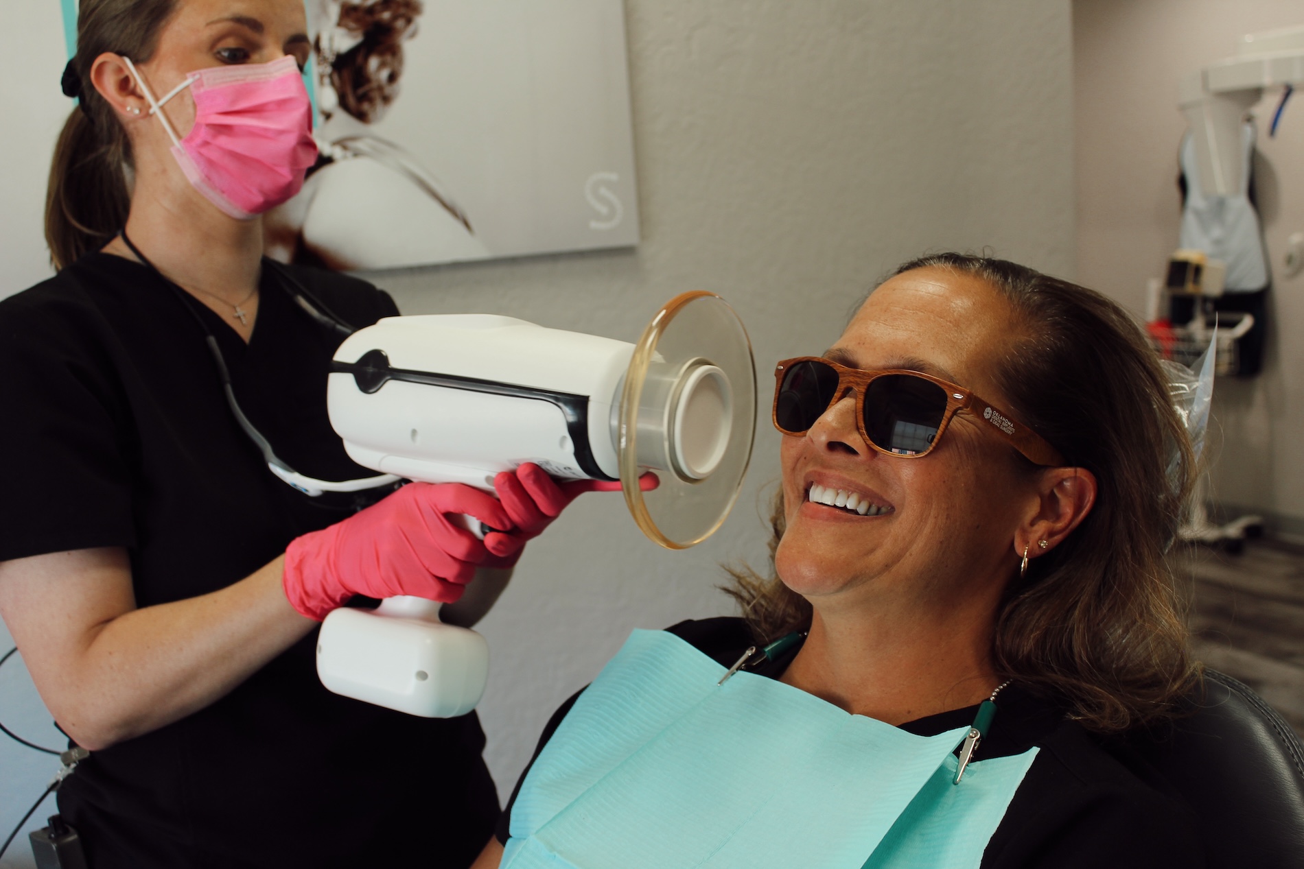 The image shows a woman receiving dental treatment, with a dental hygienist using an ultrasonic scaler on her teeth while she smiles.