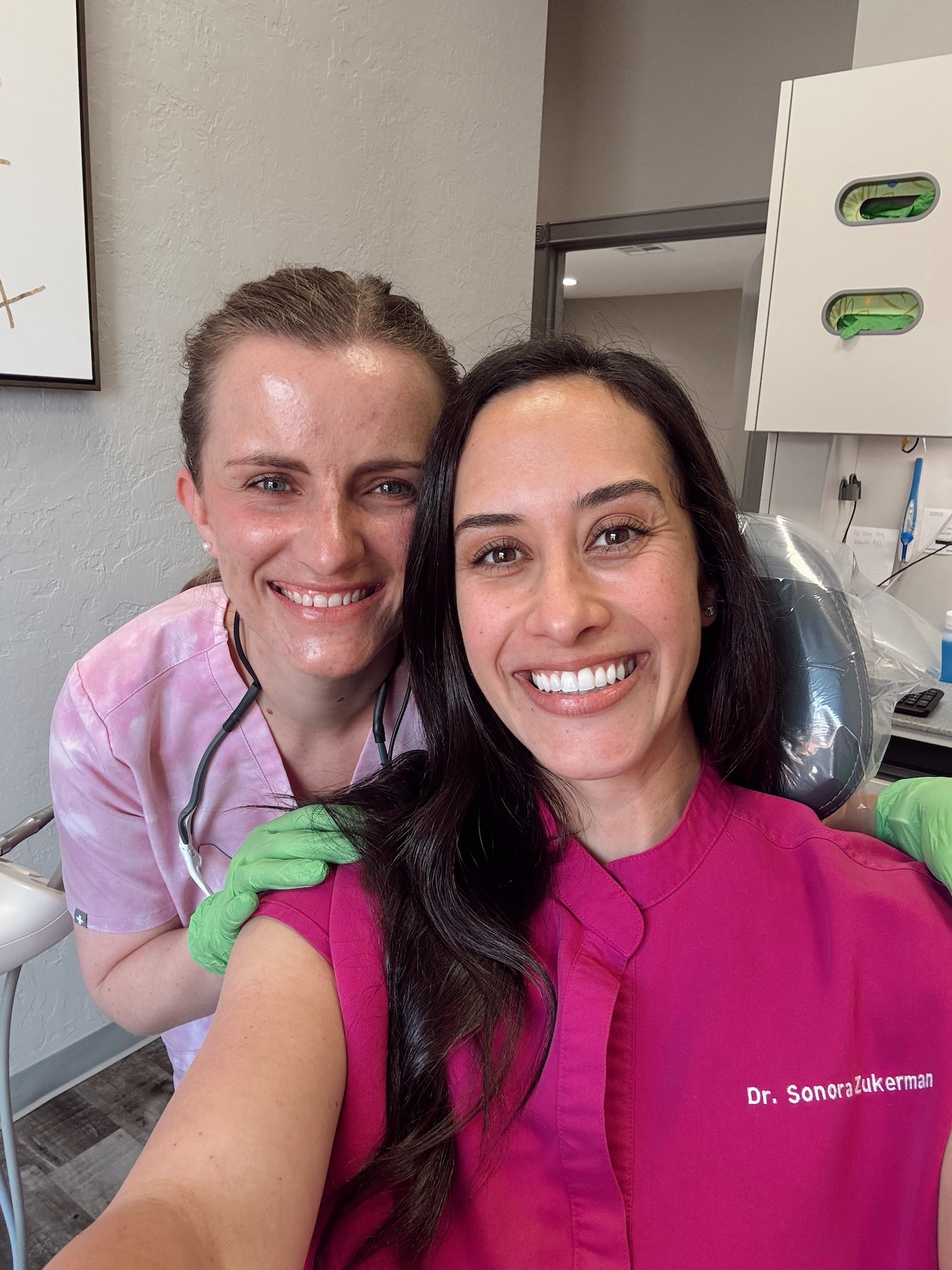 The image shows two individuals standing side by side, posing for a selfie with a smiling expression. One person is wearing a white lab coat and holding a clipboard, while the other is dressed in pink scrubs and has a dental mask draped around their neck. They are both indoors, likely within a professional setting such as a medical or dental office, indicated by the presence of equipment and the attire of the individuals.