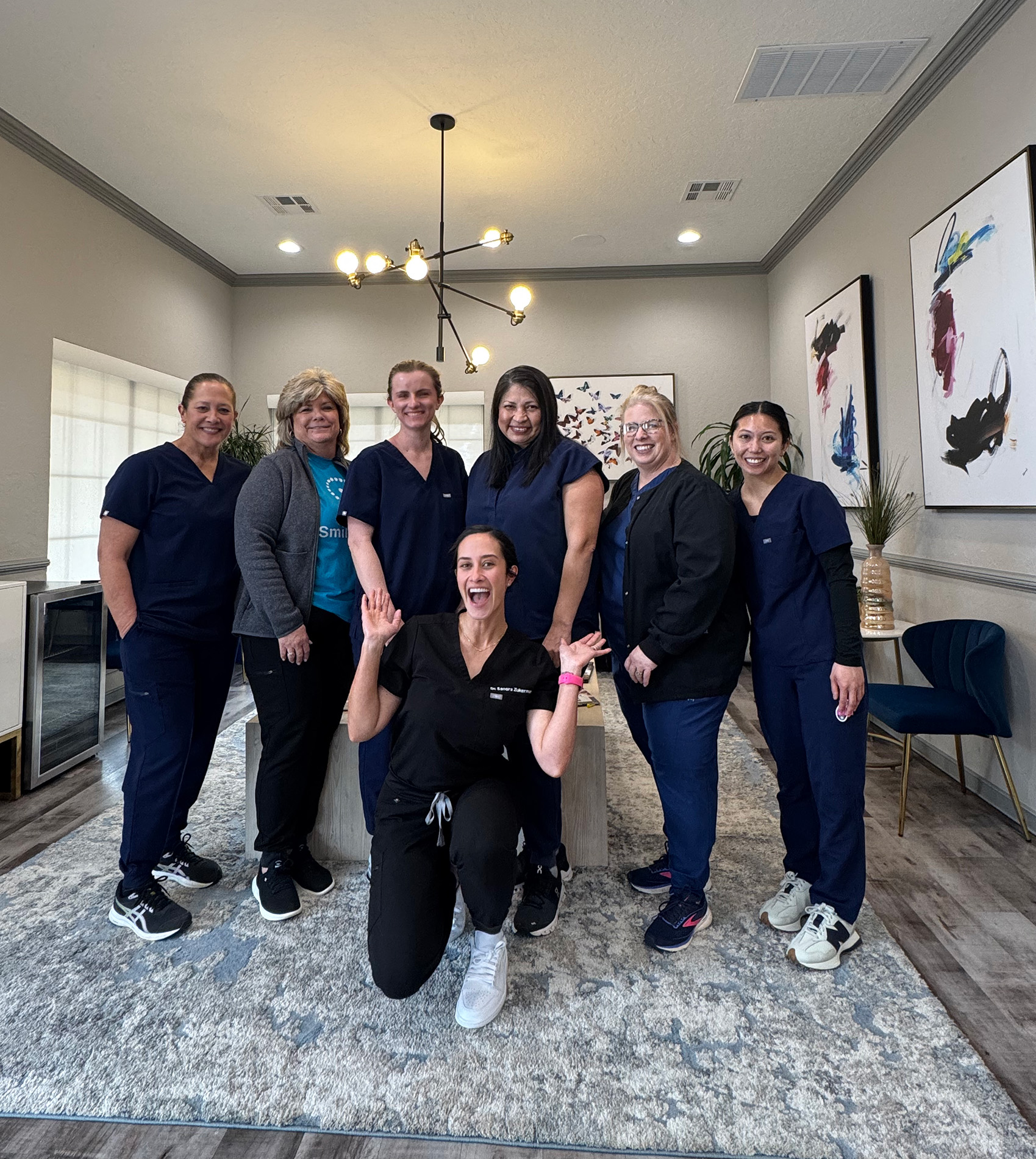 A group of six individuals posing together indoors, with a woman kneeling and smiling at the camera, standing next to a framed picture on the wall.