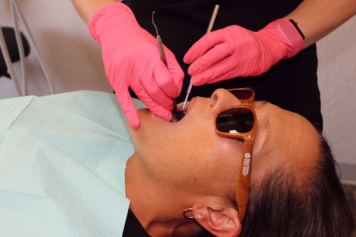 The image shows a person receiving dental care with a dental hygienist working on their teeth while wearing protective gloves and pink shoe covers, and the patient is wearing sunglasses over their eyes.