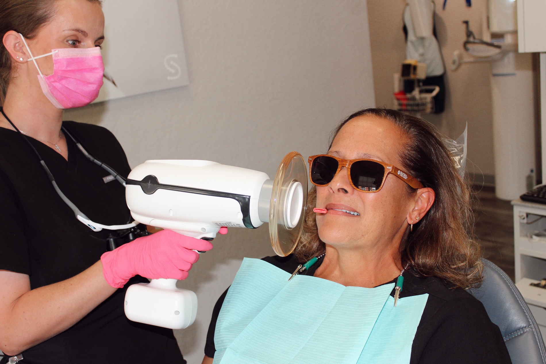 The image depicts a woman receiving dental treatment with an electronic device while a female dental professional uses a handheld device, likely for cleaning or polishing teeth, and applies a blue protective mask over the patient s face.