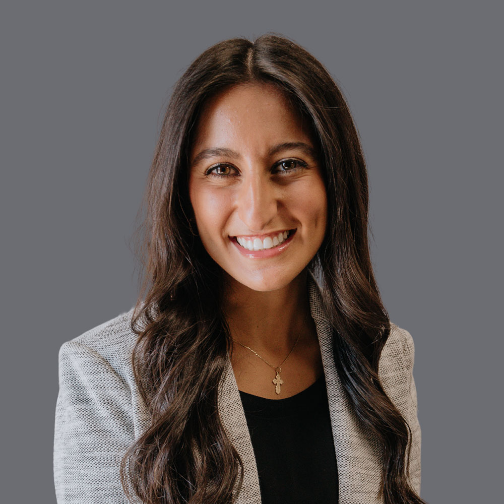 A smiling woman with long hair, wearing a light-colored jacket, poses for a professional portrait against a neutral background.