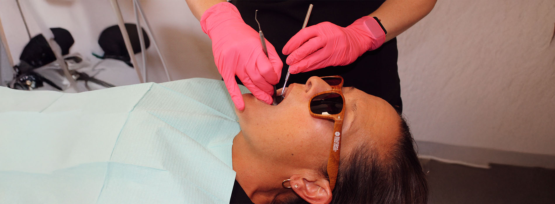 The image shows a person with sunglasses lying on a dental chair, receiving oral care from a professional who appears to be applying a substance to their teeth using a syringe.