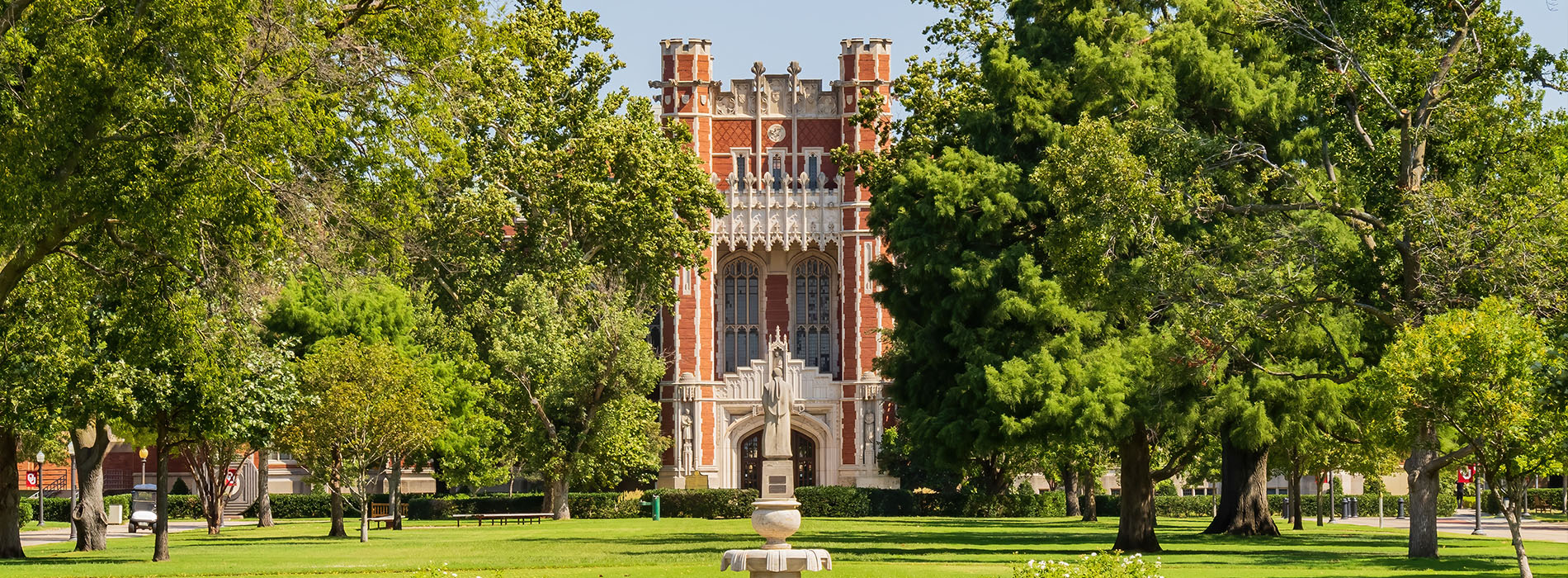 The image depicts a large, historic building with a prominent tower and clock face, surrounded by lush greenery and trees, likely part of a college campus.