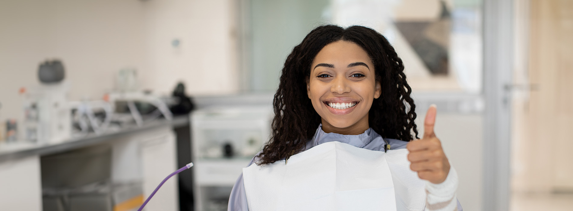 The image shows a person smiling at the camera while wearing a white lab coat, standing in front of a laboratory setting with various pieces of equipment visible in the background.