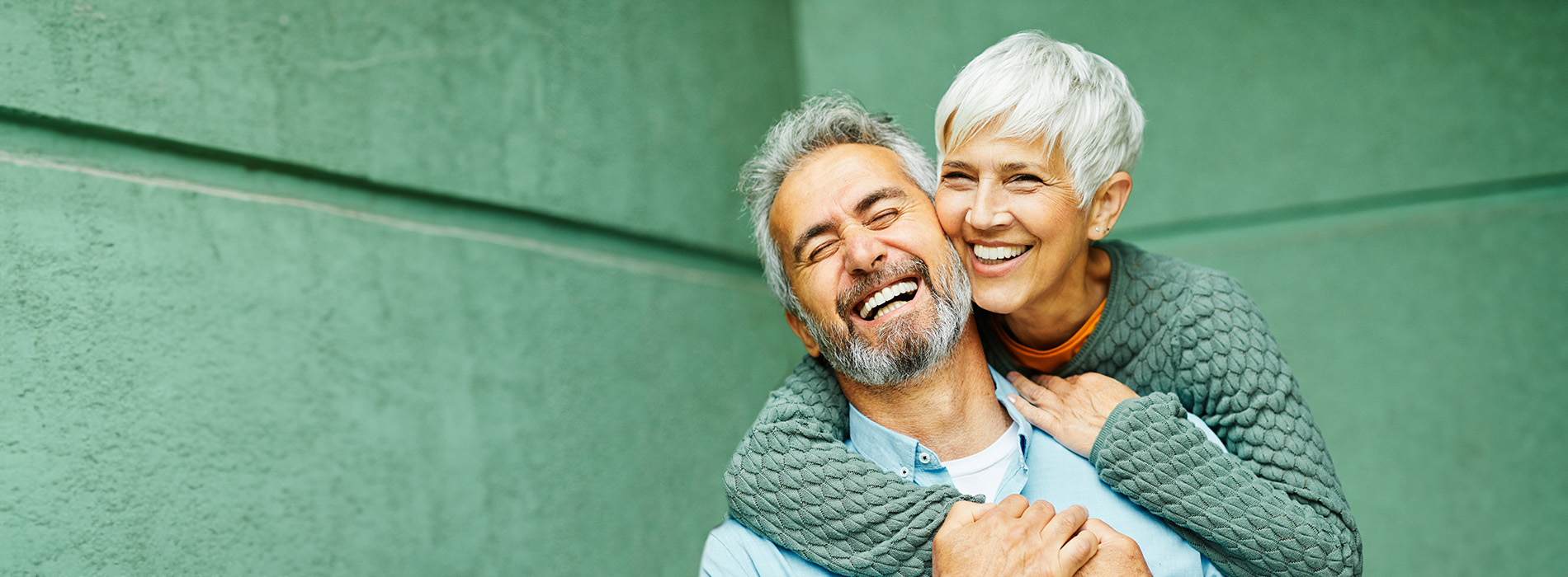A man and woman are embracing with smiles on their faces, standing outdoors against a green wall.