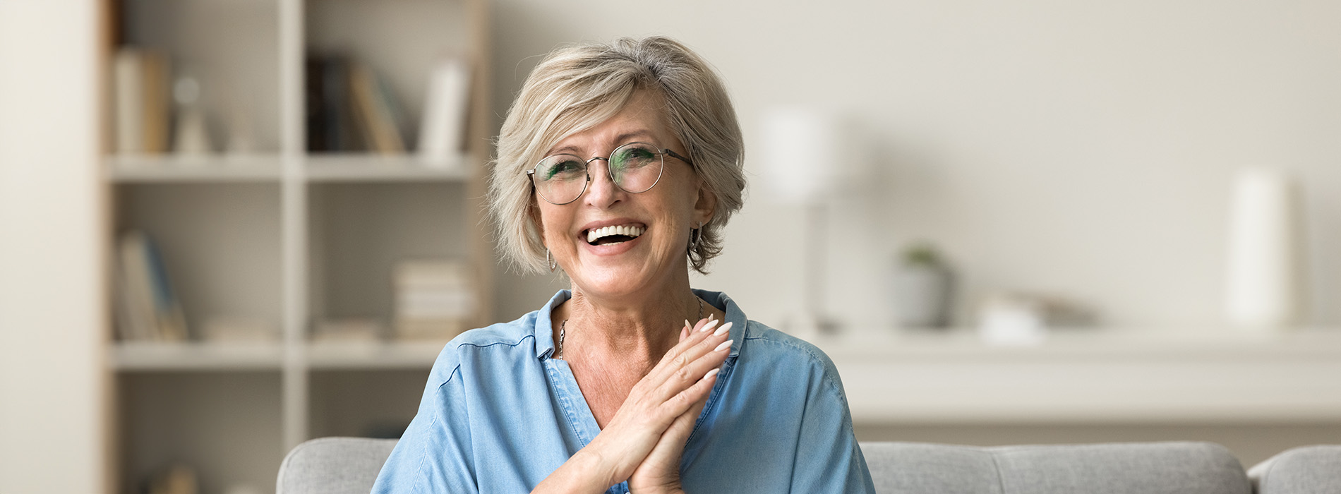 A woman with glasses is smiling, sitting on a couch with her hands clasped together.