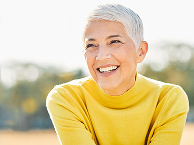 The image shows a person with short hair, wearing a yellow top, smiling at the camera against an outdoor background.