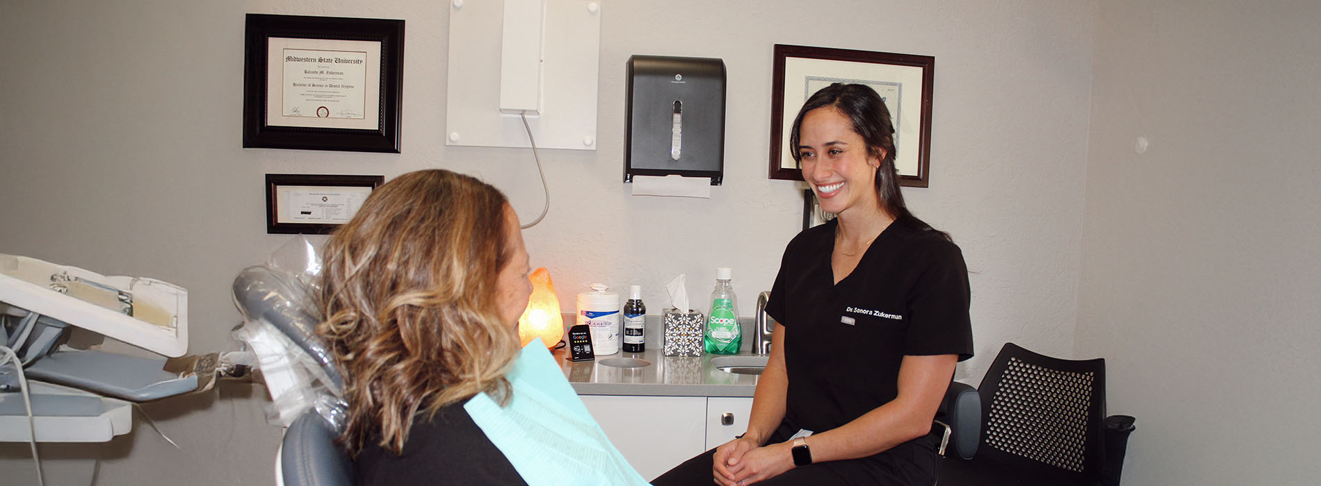 In the image, there are two individuals in a dental office setting. One person is seated in a dental chair while another stands behind them, both wearing masks. The standing individual appears to be a dental professional, possibly a dentist or hygienist, engaging with the seated patient who seems to be waiting for treatment. They are situated within an office environment that includes typical dental equipment and decor.