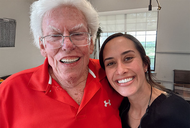 The image shows an older man with white hair wearing glasses and a red shirt, smiling at the camera next to a younger woman who is also smiling and has her arm around him. They are both indoors, and there appears to be a window in the background.