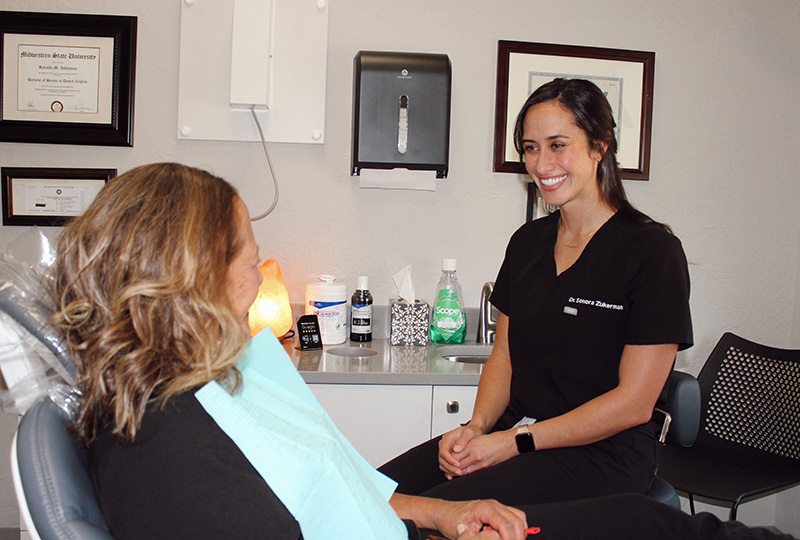 A dental professional seated at a desk with a patient, both smiling, in an office setting.