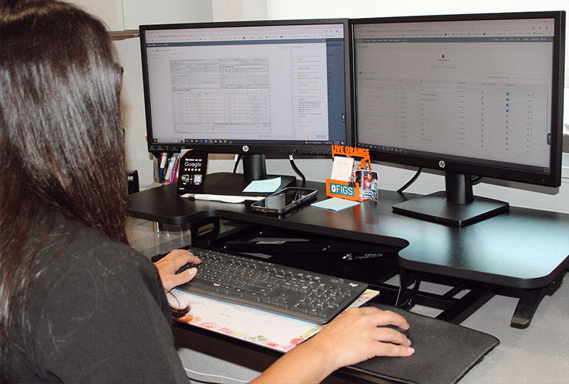 A person sitting at a desk with dual computer monitors displaying data, working on a laptop, and using a keyboard with their right hand.