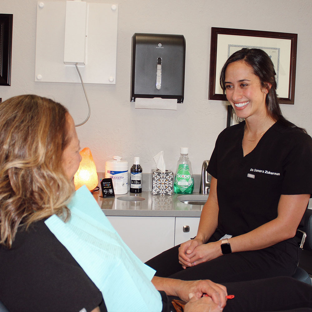 The image shows a dental hygienist seated at a desk, smiling and looking towards the camera, with a patient seated across from her, both in an office setting.