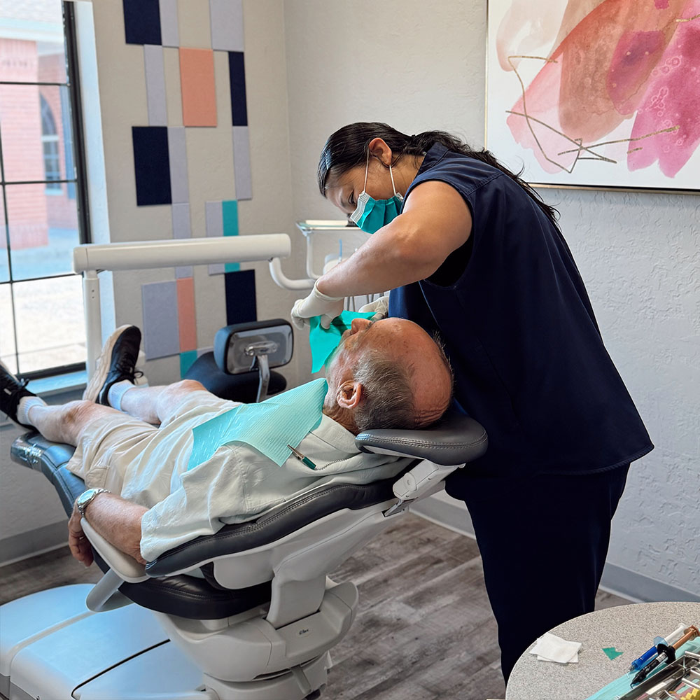 The image depicts a dental office setting with a dentist performing a procedure on a patient who is lying down on a dental chair, with a dental hygienist assisting during the process.