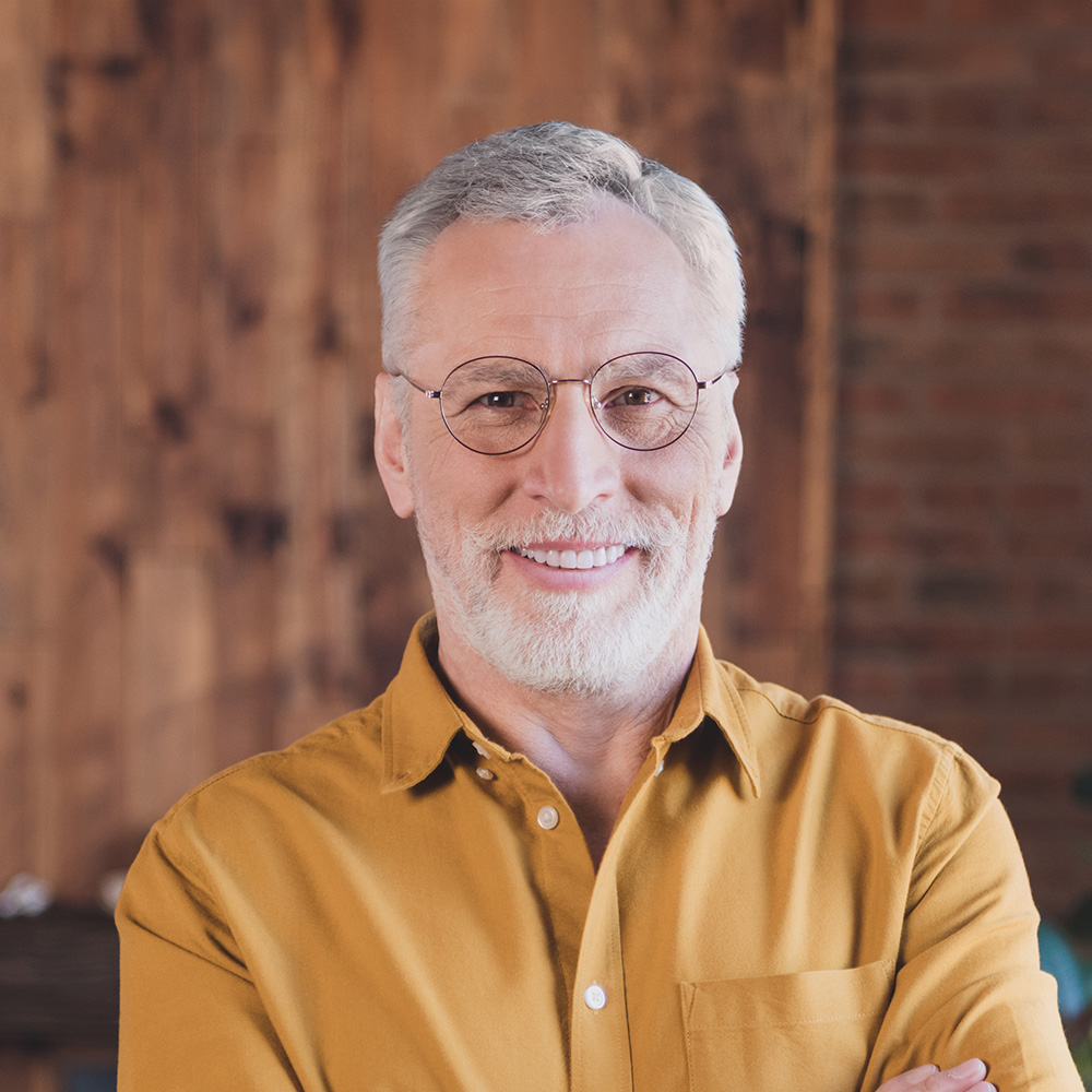 The image features an older man with white hair and glasses, wearing a yellow shirt, standing confidently with his arms crossed.