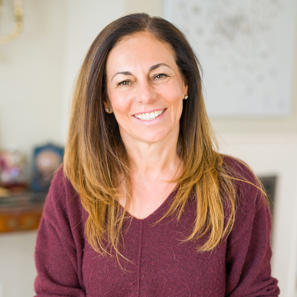 The image shows a woman with long hair smiling at the camera, wearing a dark top with lighter sleeves and a maroon cardigan over it.