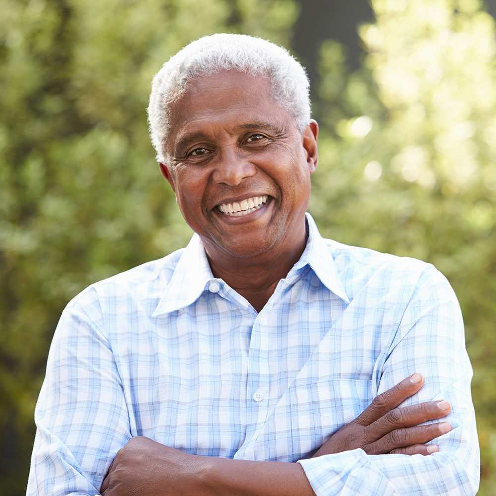 The image shows an older man with a smile, wearing a light blue shirt and standing outdoors with his arms crossed.