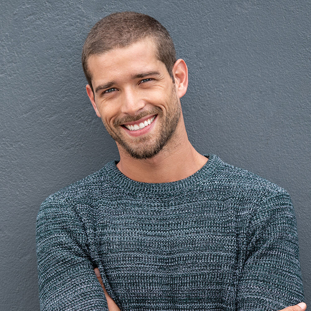 The image shows a man smiling at the camera with his arms crossed over his chest. He has short hair and is wearing a dark blue sweater, standing against a gray wall.