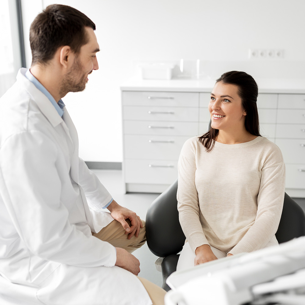 A man and a woman are seated in a dental office, with the dentist smiling at the camera while gesturing towards her patient.