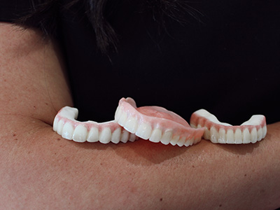 The image shows a person holding two artificial teeth with visible false gums, which are pink and appear to be made from a material that mimics natural tooth enamel.