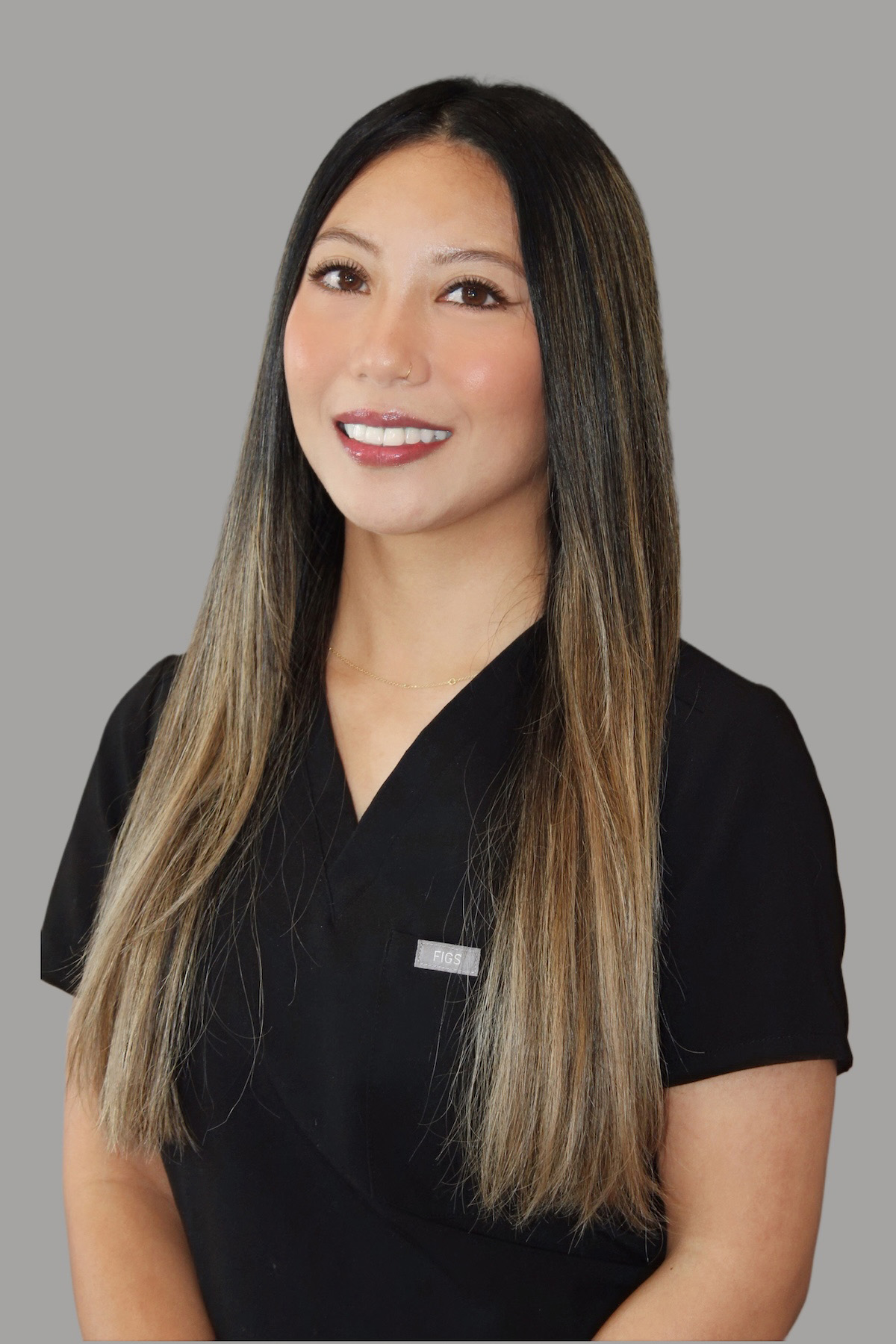 The image depicts a woman wearing a black shirt with a badge on her chest, posing for a professional headshot with a neutral background.