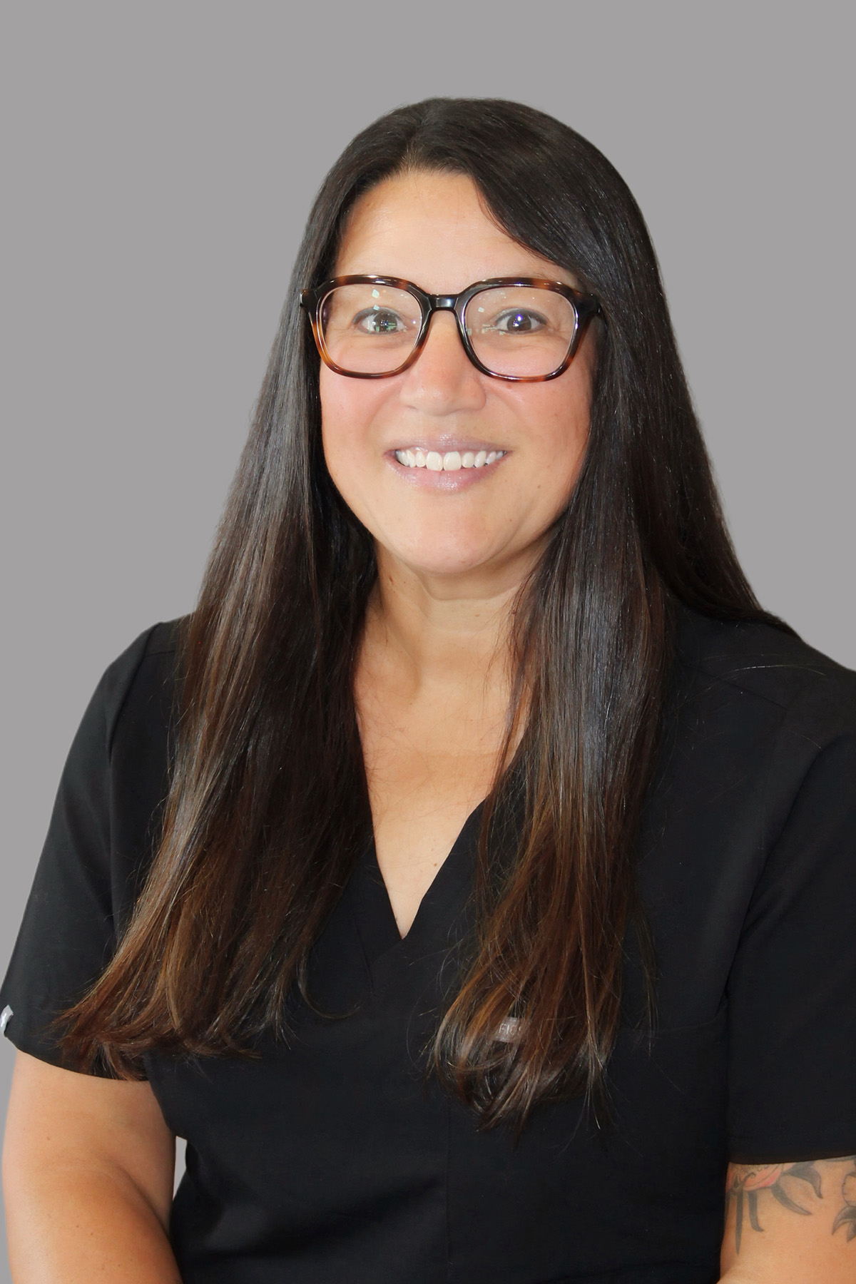 Woman with long hair wearing glasses, smiling at the camera, dressed in a dark shirt and sitting against a neutral background.