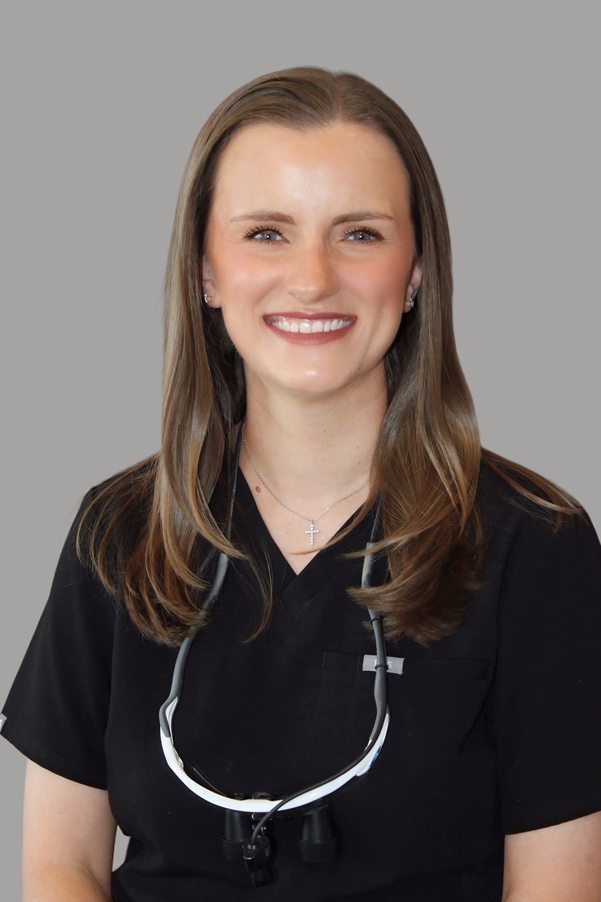 The image shows a woman wearing a black scrub top, stethoscope around her neck, and a name tag with a lanyard. She has short brown hair, is smiling at the camera, and appears to be in a professional setting.