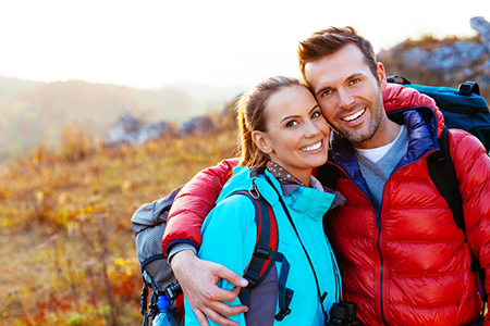 A man and woman are posing together outdoors, with the man wearing a backpack and both dressed appropriately for hiking or outdoor adventure.