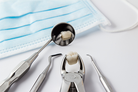 The image shows a collection of dental instruments including a pair of pliers with a tooth, placed on top of a blue surgical mask, which is resting on a white surface.