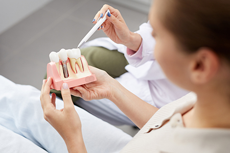 A person wearing a dental mask demonstrates a dental implant model with a handheld device.