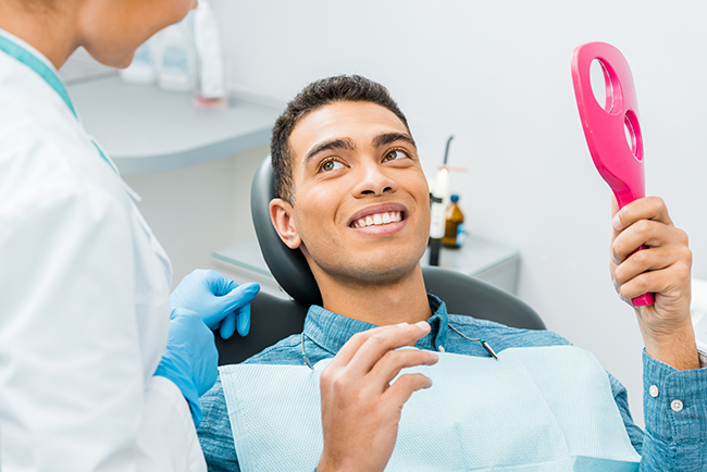 The image shows a smiling man seated in a dental chair with a pink dental device in his mouth, being attended to by a dental professional wearing protective gloves and a face mask.