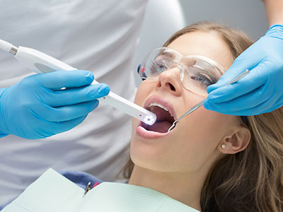 A woman sitting in a dental chair with her mouth open, receiving a dental procedure, while a dental professional uses a device to examine her teeth.